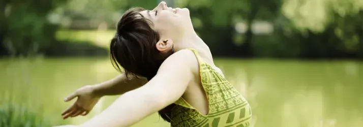 A woman outdoors with arms outstretched and eyes closed, appearing to breathe freely and enjoy nature, suggesting relief from allergies.