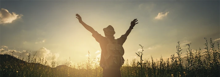 A person with a backpack standing in a field of tall flowers, raising their arms toward the sky during a golden sunset, symbolizing freedom and vitality.