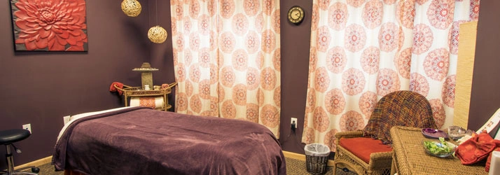 A cozy massage therapy room with a treatment table, warm lighting, patterned curtains, and calming decor at Cram Chiropractic & Wellness Center